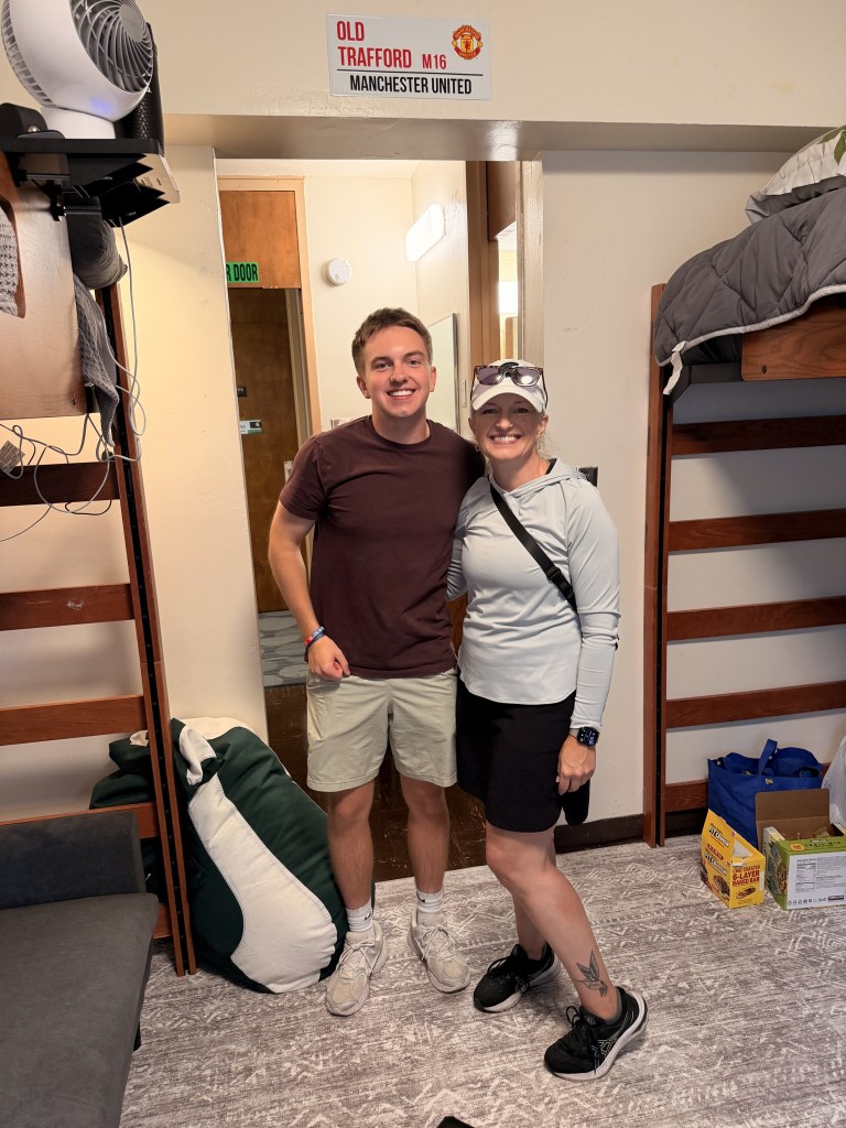 Two people (author and her son) standing in the doorway next to a lofted residence hall bed.